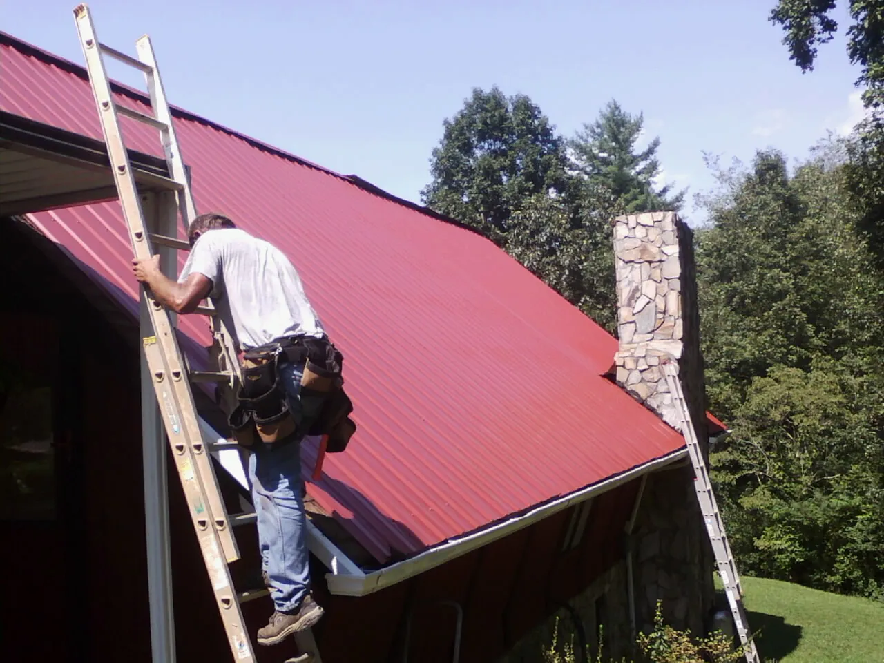 Roofer on a ladder beside a red metal roof and stone chimney — Soffit & Fascia Repair by Roofing Brothers in Farmville